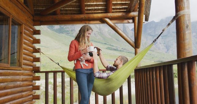 Couple relaxing in hammock on porch of rustic log cabin with stunning mountain view. Woman holding a small dog while a man sits in the hammock, enjoying the peaceful outdoor setting. Ideal for use in articles or advertisements about travel, nature retreats, romantic getaways, outdoor living, or pet-friendly vacations.