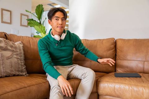 Young asian man reaching for tablet on tan leather sectional couch wearing white headphones