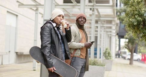 Urban skateboarder friends standing on sidewalk drinking coffee and checking smartphone