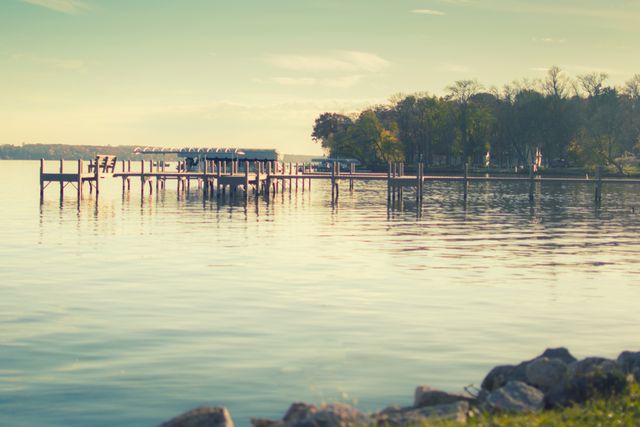 Calm lakefront scene with a long dock extending over the placid water. Trees line the shore, adding to the serene, peaceful atmosphere. Rocks in the foreground add rustic texture. Ideal for use in travel promotions, relaxation or meditation media, nature websites, and outdoor activity brochures.