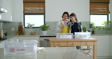 Diverse female friends sorting recycling in modern kitchen with paper and plastic bins