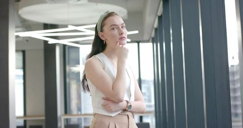 Pensive professional woman standing by window in modern coworking space, minimalist design