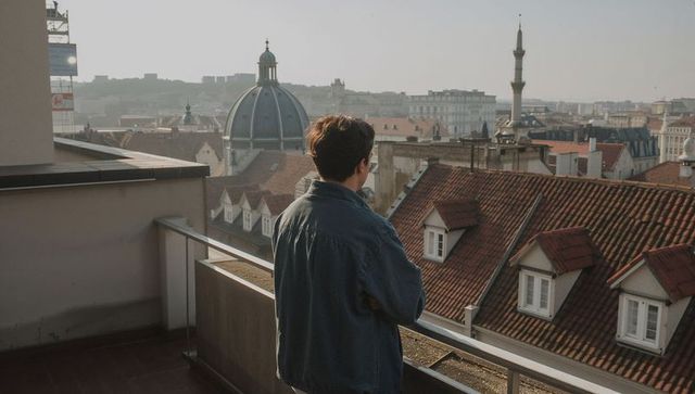 Young man leaning on rooftop railing viewing historic european red tile roofs and dome