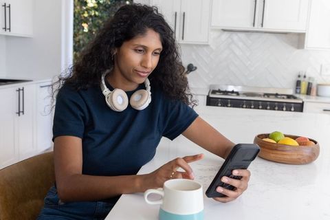 African american woman wearing headphones checking smartphone at bright kitchen island