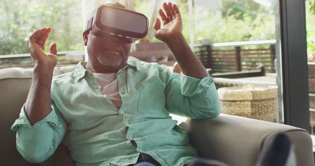 Senior man wearing a virtual reality headset and having an immersive experience on his sofa at home. Ideal for use in articles and promotions about technology for seniors, virtual reality gaming, home entertainment, and innovations in tech.
