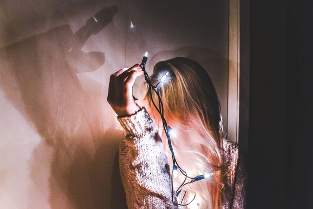 Blonde woman holding string lights close to her hair in a dimly lit room, creating cozy and mysterious ambiance. Suitable for themes of home decoration, introspective moments, winter holidays, creative lighting ideas, and personal reflection.