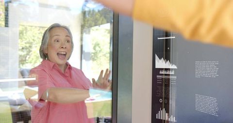 Excited senior Asian woman interacting with smart glass panel displaying digital HUD charts