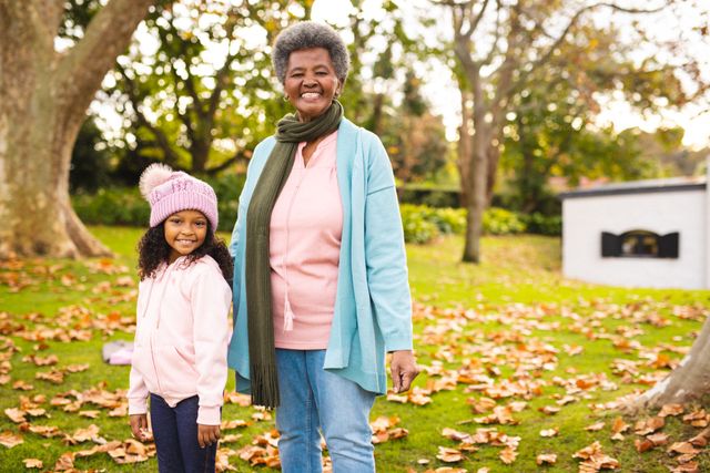 This image captures a joyful moment between a grandmother and her granddaughter standing in a park during autumn. The grandmother is wearing a scarf and light blue cardigan, while the granddaughter is dressed in a pink hat and hoodie. Ideal for use in family-oriented content, advertisements promoting outdoor activities, or articles about intergenerational relationships and bonding.