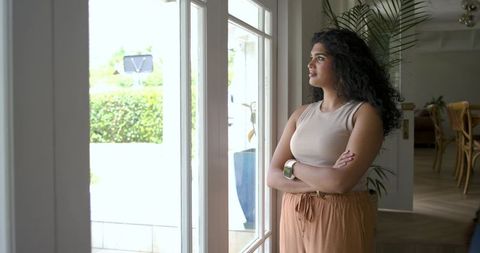 Serene indian woman standing by glass doors looking outside wearing smartwatch in home