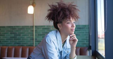 Contemplative young woman gazing out window in cafe, leaning on table in denim jacket