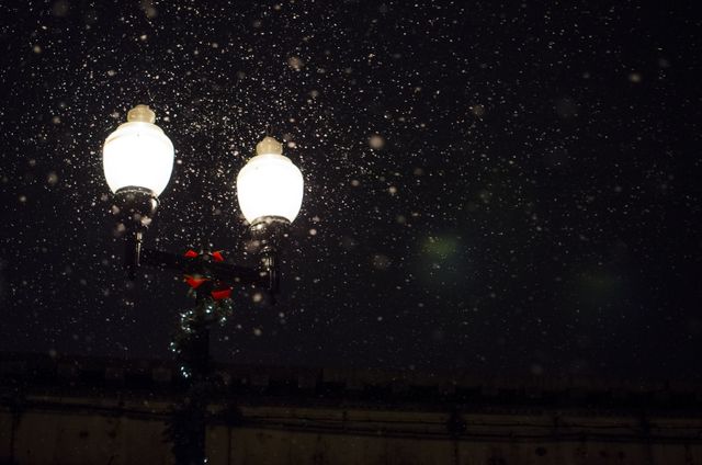 Snow falling under a street lamp creates a tranquil winter night scene. This captures the serene beauty of snowfall in an urban setting, perfect for use in winter or holiday-themed articles, advertisements, and decorations.
