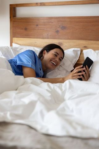Smiling woman relaxing in blue pajamas on white bed holding smartphone morning routine