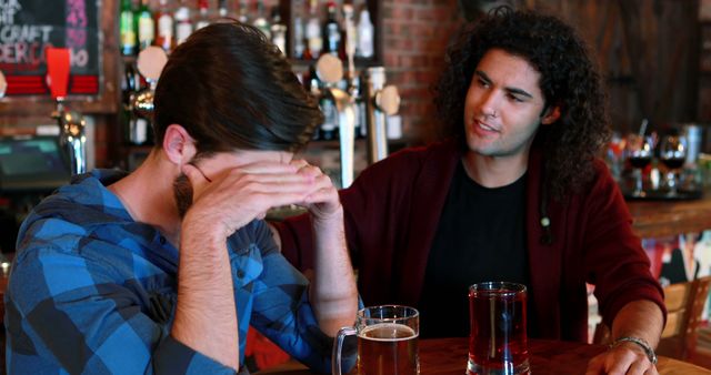 Two friends sitting at a bar, engaged in a conversation. One of them looks stressed and is covering his face with his hands, while the other is offering support. Background features various bottles and bar decor. Suitable for themes related to friendship, emotional support, social interactions, and stress relief.