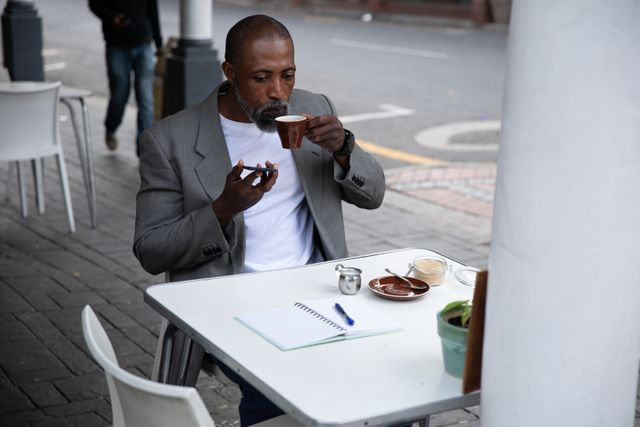 Smart casually dressed African American man with short hair and a grey beard out and about in the city during the day sitting at a table outside a cafe by enjoying a cup of coffee holding smartphone