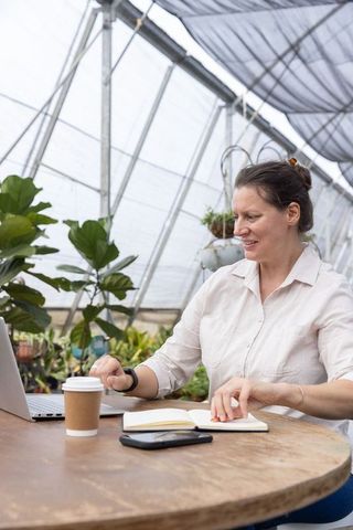 Woman working in greenhouse with laptop and notebook at round wooden table among plants