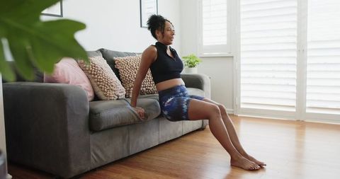 African american woman performing triceps dips on sofa edge in bright living room