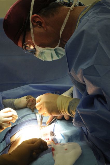 Surgeon is performing a medical procedure in an operating room. He is wearing scrubs and a face mask, exuding focus and precision. Gloved hands of assistant visible helping with the surgery. Ideal for use in healthcare-related articles, medical journals, and educational materials to depict surgical procedures or highlight the medical profession.