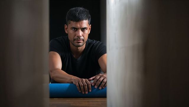 Focused man leaning on blue yoga mat between pillars in dim fitness studio moody portrait