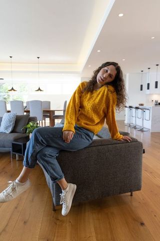 Young woman sitting on sofa back wearing mustard sweater in modern open-plan living room