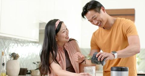 Diverse couple blending green smoothie in modern kitchen, smiling and sharing