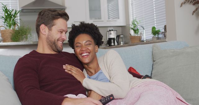 Interracial couple enjoying a relaxed moment at home, sitting on the couch and laughing together. Useful for content depicting love, relationships, domestic happiness, and interracial harmony. Ideal for lifestyle, family life, and home decor articles or promotions.