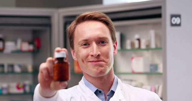 A young Caucasian male pharmacist presents a bottle of medicine, with copy space. His professional attire and the pharmacy setting emphasize the importance of trusted healthcare providers.