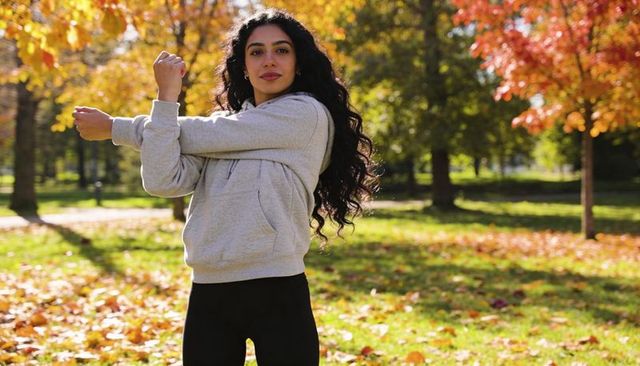 Young woman stretching arm across chest in autumn park sunlight wearing hoodie and leggings