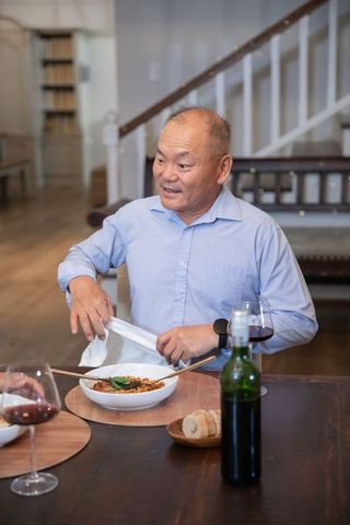 Mature chinese man enjoying cozy home dinner holding napkin beside wine and pasta bowl