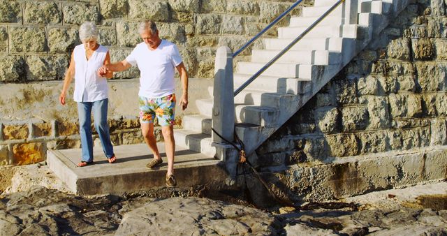 Happy senior caucasian couple holding hands and walking from stairs to the beach by sea. Retirement, relationship, love, togetherness, free time and relaxing, unaltered.