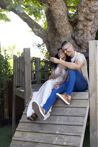Couple sitting on wooden ramp sharing white daisy under treehouse, smiling romantic moment