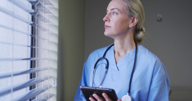 Nurse in medical uniform holding digital tablet and gazing outside window, conveying thoughtful emotion. Ideal for use in medical and healthcare ads, brochures illustrating modern healthcare technologies, and articles highlighting nursing roles and responsibilities.