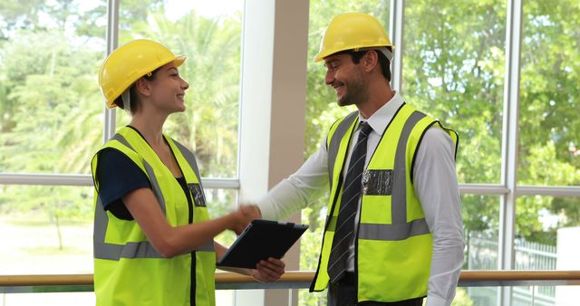 Male and female construction engineers wearing yellow safety vests and hard hats, shaking hands and smiling in front of large windows. This photo can be used for demonstrating teamwork, professional agreements, workplace safety, and cooperation within engineering and construction industries.