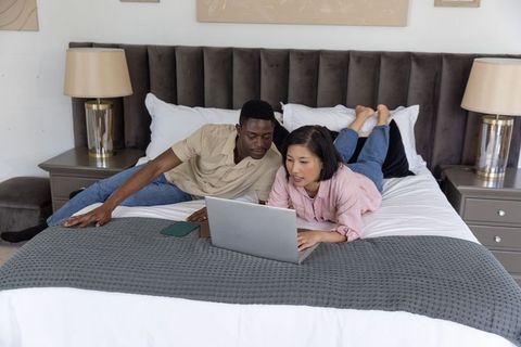 Diverse couple lying on bed browsing laptop in cozy neutral bedroom planning together