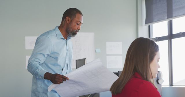 A male architect carefully reviewing building plans while a female colleague works at a desk in a modern office with natural light. Useful for depicting industry professions, teamwork, and design projects in a contemporary workspace. Ideal for content related to architecture, engineering, business collaboration, and professional environments.