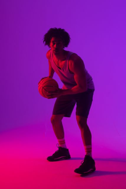 Vertical image of african american male basketball player holding ball, in purple light, copy space. Athlete, sport, competition, confidence and fitness concept.