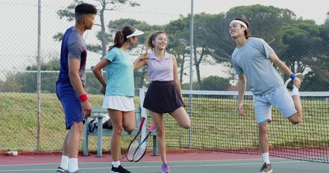 Diverse friends stretching and laughing on outdoor tennis court before doubles match