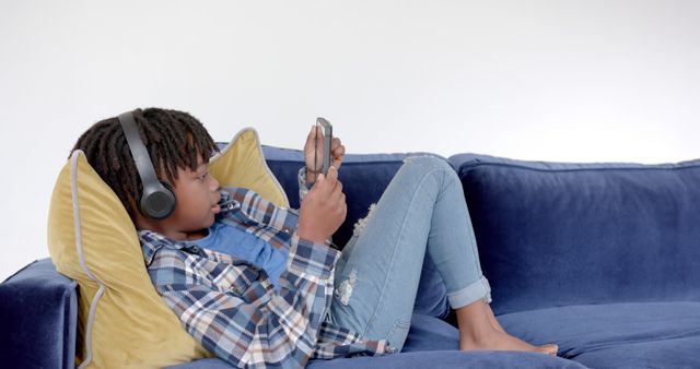 Black child lying on a blue couch, wearing headphones and focused on playing video games. The casual and relaxed scene is perfect for illustrating technology use among children, casual entertainment, or home leisure activities.