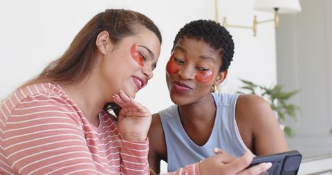 Two friends enjoying self-care with under-eye patches and smartphone in cozy livingroom