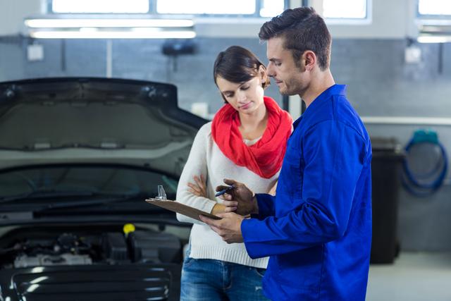 Mechanic in blue uniform discussing car repair checklist with female customer in a repair garage. Ideal for use in automotive service advertisements, car maintenance guides, and customer service training materials.