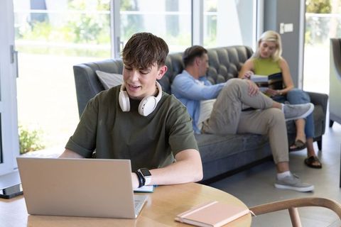 Teenager studying on laptop with headphones at home while parents relax on sofa