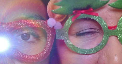 Festive sparkle closeup of two women wearing glitter novelty glasses with lens flare, pom-poms
