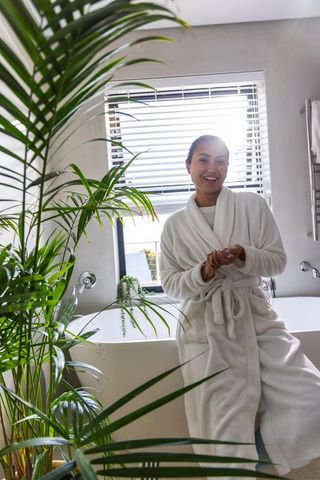 Asian woman wearing white bathrobe enjoying bright spa bathroom with lush green plants