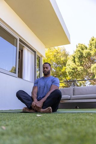 African american man meditating on terrace barefoot wearing blue shirt for mindfulness