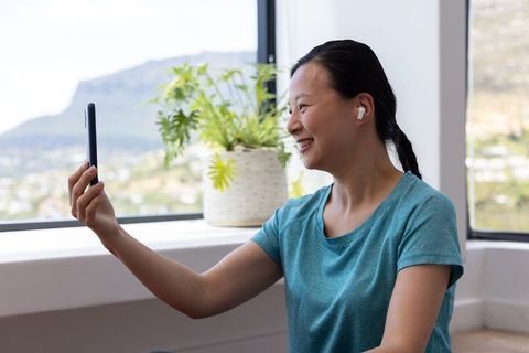 Asian woman smiling and taking video call with smartphone and earbuds near window with potted plant