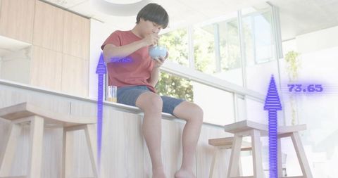 Asian man eating breakfast on kitchen counter, sitting barefoot in modern bright interior