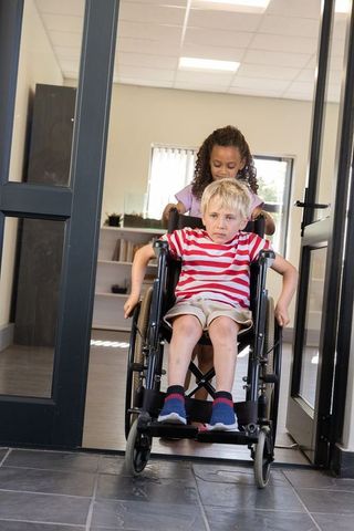 Diverse children navigating wheelchair through school doorway, girl assisting boy moving forward