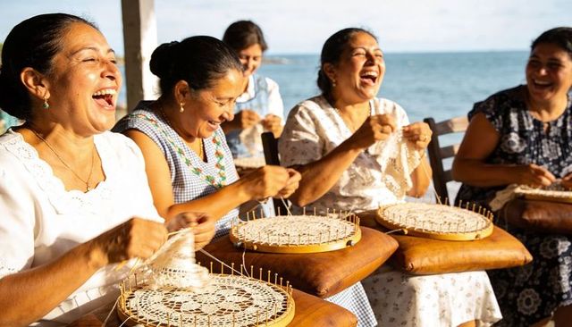 Latina women laughing and making bobbin lace on coastal veranda with wooden pillows