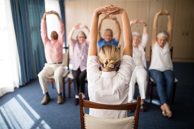 This image shows a group of senior individuals participating in a stretching exercise led by an instructor in a retirement home. It is ideal for use in materials promoting senior health, fitness programs, retirement home activities, and community wellness initiatives.