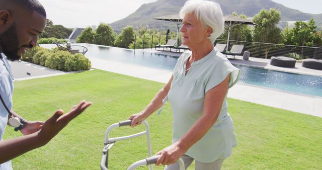 Elderly woman using walker with caregiver outdoors, standing near pool and mountainous backdrop. Ideal for images about elder care, rehabilitation, health support, active aging, senior lifestyle, physical therapy. Perfect for healthcare, senior care facilities, wellness programs.