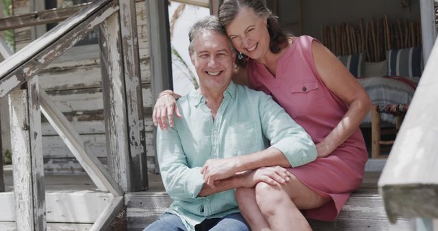 A joyful mature couple sitting together on a rustic porch, enjoying each other's company. The man wears a light green shirt while the woman is in a pink dress. This image can be used for themes such as retirement, vacation, relationships, love, and outdoor relaxation.